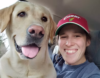 Smiling white lab sitting close to female dog sitter with red hat